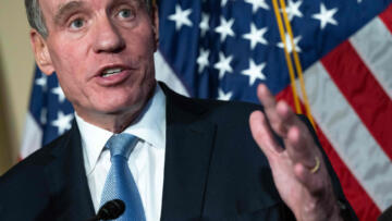 U.S. Sen. Mark Warner standing at a podium with his left hand raised. In the background, there is an American flag.