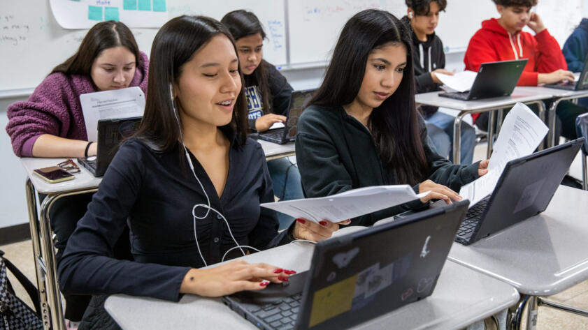 Photograph of 6 high school students on their laptop. Four of the students have long hair, two have short hair. One student has one earbud in their ear.