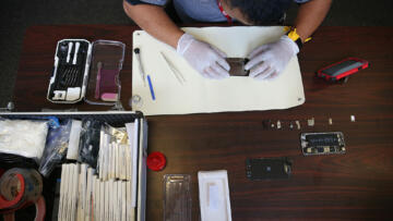 Photograph of an Asian man wearing gloves working on a phone that is taken apart. The photograph is taken from above.