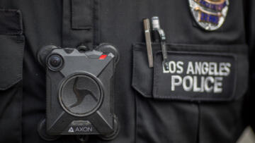 Closeup photograph of a black Axon body camera worn by a police officer. The badge next to it says “LOS ANGELES POLICE.”