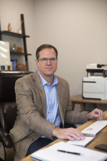 Photograph of a man wearing glasses and a plaid blazer at his desk.