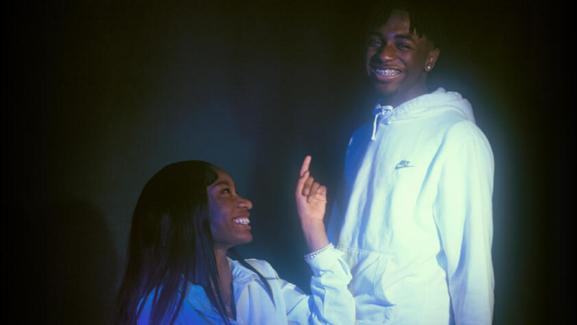 Photo of Mia Townsend and Maurice Newton, two young Black teenagers. Mia, seated and wearing a white collared shirt, is smiling and pointing at Maurice, who is wearing a white Nike hoodie.